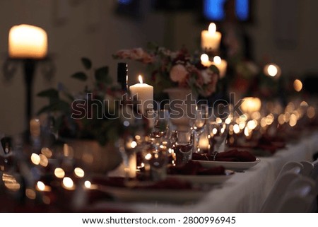 Romantic wedding reception long table setting illuminated by warm candlelight with glowing white pillars on candelabra, wine glasses, plates, maroon napkins and floral centerpiece at night