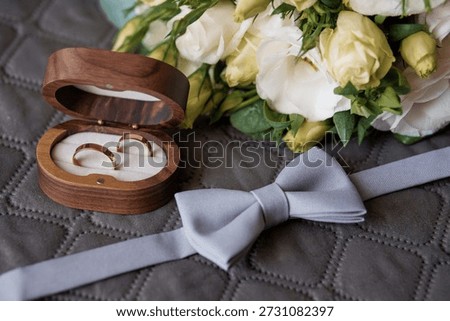 A romantic wedding setup featuring gold rings in a wooden box, white roses, and a gray bow tie on textured fabric, evoking a classic and intimate bridal theme