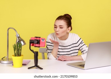 Romantic woman blogger in striped shirt recording video for followers, air sending kissing over palms hands, expressing love. Indoor studio studio shot isolated on yellow background