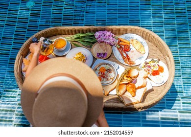 Romantic Woman In Straw Hat On Vacation Pouring Black Tea From Glass Kettle To White Ceramic Cup, Enjoying Floating Breakfast Or Lunch In Sunlit Swimming Pool In Summer, On Holidays At Resort Or Hotel