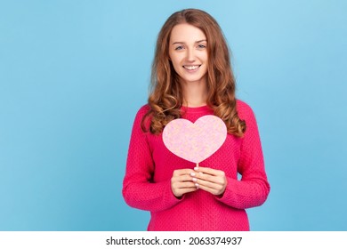 Romantic woman wearing pink pullover, holding heart symbol on stick and smiling to camera, congratulating on Valentines Day, saying I love you. Indoor studio shot isolated on blue background.