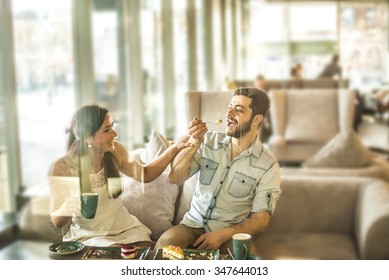 Romantic Young Adult Couple Drinking Coffee With Handsome Man Laughingly Feeding A Biscuit To His Girl Friend Or Wife Cute Woman Look At Eyes Face To Face Against Window View Sweet Food On Spoon