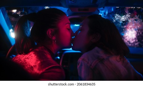 Romantic Young Adult Lesbian Female Couple Kiss On The Front Seat Of Their Car On A Rainy Night In The City. Women In Love On A Date In Neon Urban Environment. Concept Of LGBT, Relations And Driving.