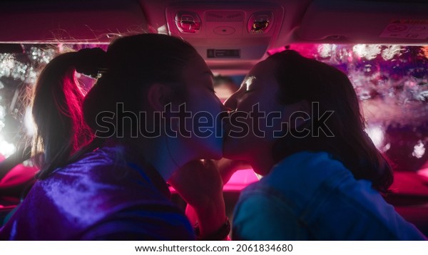 Romantic Young Adult Lesbian Female Couple Kiss\
on the Front Seat of Their Car on a Rainy Night in the City. Women\
in Love on a Date in Neon Urban Environment. Concept of LGBT,\
Relations and Driving.