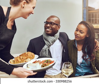 Romantic Young African American Couple Sitting Arm In Arm Being Served Dinner By A Waitress As They Sit At A Restaurant Table