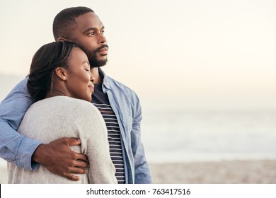 Romantic young African couple enjoying a late afternoon together on a sandy beach at sunset while standing in each other's arms 