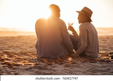 Romantic young African couple sitting together on a sandy beach at dusk toasting each other with wine  