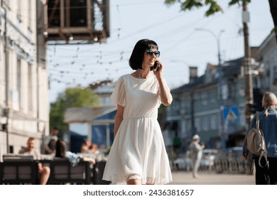 Romantic young brunette woman with short hair wears mini white dress and sunglasses, talking on cellphone, walking on a crowded city street in the summertime. Girl communicates by smartphone outdoors. - Powered by Shutterstock