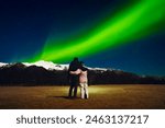 Romantic young caucasian couple together hug each other in cold night stand watch northern lights over snowy peaks in Iceland. Holiday vacation trip in autumn