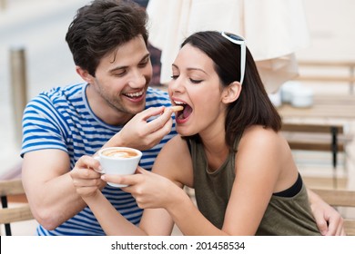 Romantic young couple drinking coffee with the young man laughingly feeding a biscuit to his girlfriend or wife as she holds a cup of cappuccino
