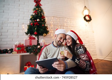 Romantic Young Couple Enjoying Christmas Time With A Cup Of Tea And Tablet At Home.