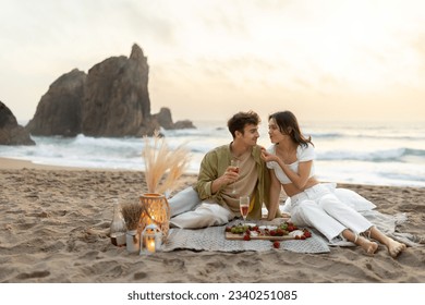 Romantic young couple having picnic on sandy beach with ocean view, drinking wine and eating fruits, enjoying date in the evening at coastline, free space - Powered by Shutterstock