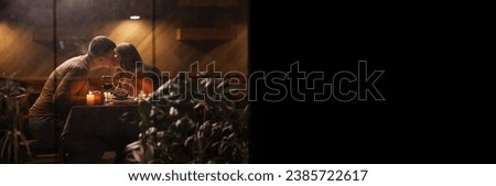 Romantic young couple kiss while during dinner at dining table celebrating Valentine's Day, view through window. Banner. Copy space