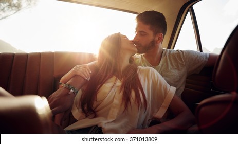 Romantic young couple sitting in back seat of a car and kissing . Affectionate couple on rear seat of a vehicle with sun flare.