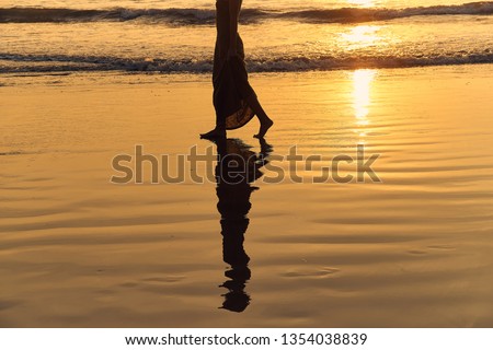 Romantic young girl walks on the beach barefoot in the water. Woman stroll barefoot in the sea at sunset. Beautiful female legs in the ocean. Silhouette of a female body in a dress on sea background.