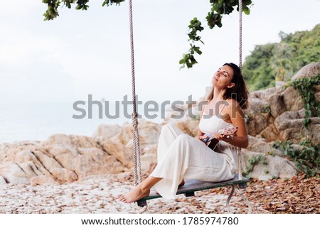 Romantic young happy calm caucasian tattooed lonely woman with ukulele on swing tropical rocky beach at sunset.	 