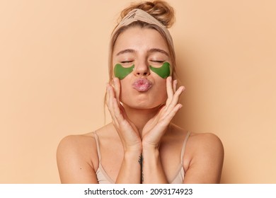 Romantic Young Woman Keeps Eyes Closed Lips Rounded Touches Face Gently Applies Green Collagen Patches Undergoes Beauty Treatments Wears Headband Rejuvenates Skin Isolated Over Brown Background