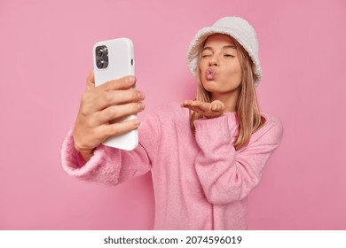 Romantic young woman sends air kiss at cellular camera takes flirty selfie photo winks eye and pouts lips wears hat and jumper isolated over pink background enjoys video chat with boyfriend.