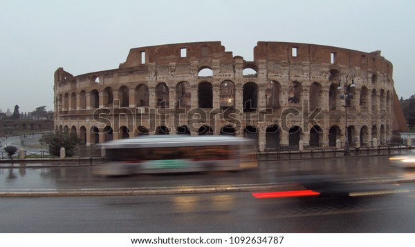 Rome, Coliseum view in a Rainy \
Dusk, cars and bus with light on reflecting in wet\
asphalt