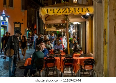 Rome, Italy - June 4, 2018: People Dine And Mingle At A Sidewalk Pizzeria At Night On A Busy Street In Rome. 