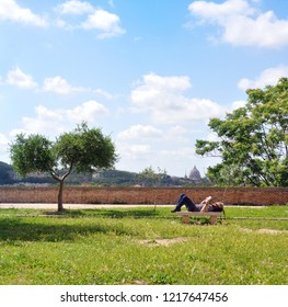 Rome, Italy - May 13 2018: St Pietro In The Skyline. Giardino Degli Aranci (the Orange Garden) Or Otherwise Known As Parco Savello. Man Resting And Reading A Book. 