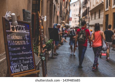 ROME / ITALY - May 26, 2018 - Typical Narrow And Busy Street In Rome, Italy With A Restaurant Board With A Pizza Menu And Pedestrians Walking On The Background