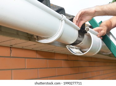 Roof Gutter Installation And Repair. A Man On A Ladder Is Replacing A Plastic Rain Gutter Joint, Bracket, Connecting The Gutters Together.