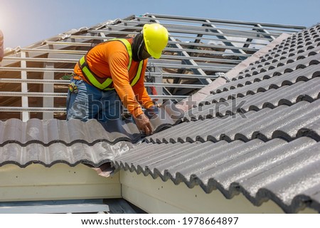 Roof repair, worker with white gloves replacing gray tiles or shingles on house with blue sky as background and copy space, Roofing - construction worker standing on a roof covering it with tiles.