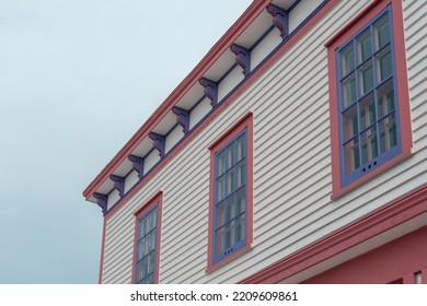 The Roof Section Of A Large White Vintage Wooden House With Decorative Pink And Purple Wood Trim. There Are Three Multi-pane Windows On The Top Floor. The Background Is A Dramatic Blue Cloudy Sky.