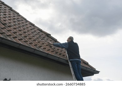 Roofer cleans a gutter with a cleaning tool while standing on a ladder, performing the necessary maintenance to prevent roof leaks and ensure proper drainage of rainwater