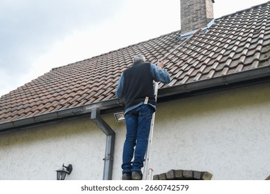 Roofer cleans gutter with cleaning tool while standing on ladder, performing necessary maintenance to prevent roof leaks and ensure proper drainage of rainwater