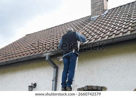 Roofer cleans gutter with cleaning tool while standing on ladder, performing necessary maintenance to prevent roof leaks and ensure proper drainage of rainwater