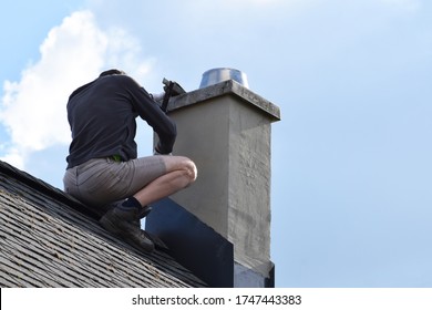 Roofer Construction Worker Repairing Chimney On Grey Slate Shingles Roof Of Domestic House, Blue Sky Background With Copy Space.
