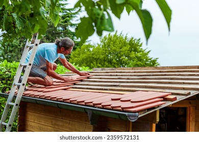 Roofer at work, installing clay roof tiles. back garden with shed, summer house garden timber outbuilding. new roof of the shed in the back yard. install tile on roof of old house.  - Powered by Shutterstock