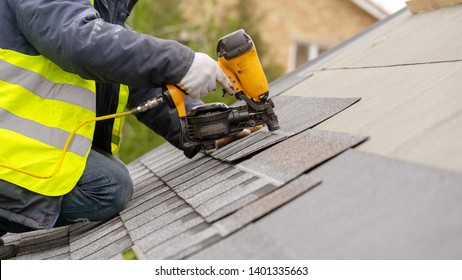 Roofer worker in special protective work wear and gloves, using air or pneumatic nail gun and installing asphalt or bitumen shingle on top of the new roof under construction residential building