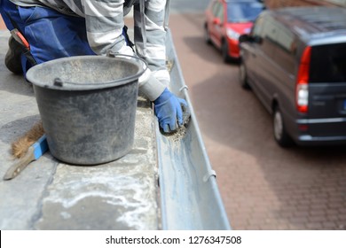 Roofer Working On Roof Maintenance In March. Clean Zinc Gutter With Blue Working Gloves