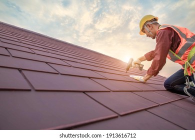Roofer working in special protective work wear gloves, using air or pneumatic nail gun installing concrete or CPAC cement roofing tiles on top of the new roof under construction residential building - Shutterstock ID 1913616712