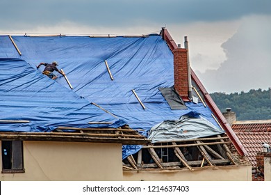 The Roofer Works On Roof When Is Rain. The Tarp Covers The Roof Of The Old House In The Reconstruction.