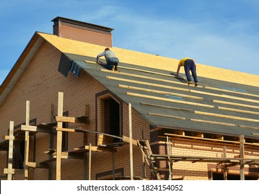 Roofing contractors are installing underlayment, waterproofing membrane on a roof deck, plywood sheathing of a large brick house under construction with scaffolding. - Shutterstock ID 1824154052