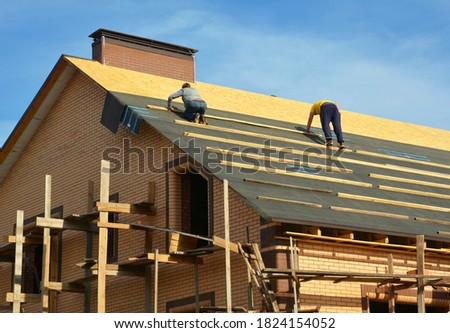 Roofing contractors are installing underlayment, waterproofing membrane on a roof deck, plywood sheathing of a large brick house under construction with scaffolding.