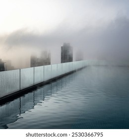 A rooftop infinity pool is shrouded in thick fog, with only the faint outlines of buildings visible through the mist. The foggy atmosphere blends with the water’s surface, creating a tranquil. - Powered by Shutterstock