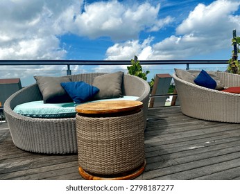 Rooftop Outdoor Lounge With Wicker Sofas, Cushions and Wooden Side Table Under Blue Sky. 

A stylish rooftop outdoor lounge featuring round wicker sofas with colorful cushions and a wooden side table beneath a bright blue sky. The open-air terrace setting overlooks greenery and creates a relaxing atmosphere ideal for hospitality, leisure and contemporary outdoor living.