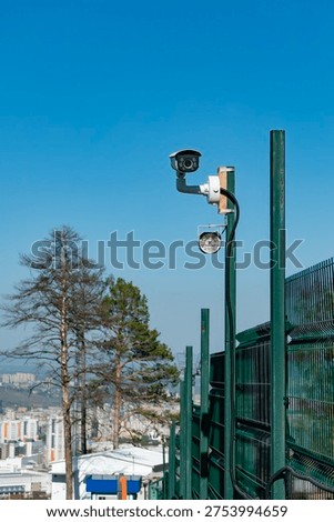 A rooftop security camera mounted on a green perimeter fence monitors an urban cityscape, conveying surveillance, safety and vigilance with cables and a floodlight against a clear blue sky.