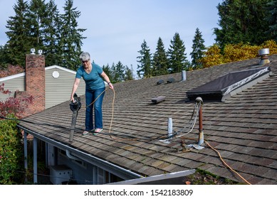 Rooftop View Of Suburban Home, Mature Woman With Leaf Blower Cleaning Roof And Gutter
