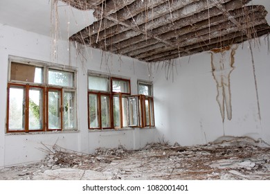 Room In Abandoned Building With Broken Windows, Destroyed Floorboards Full Of Rubble, Walls With Moisture Patches And Hanging Ceiling Insulation