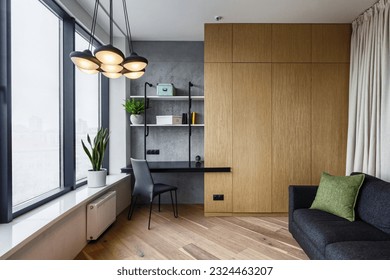 room with wooden wardrobe and cushion on grey comfort couch. home office with desk and chair near window. concept of modern interior in stylish apartment