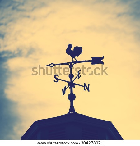 Rooster weather vane on a rooftop with an arrow and North-South pointer to show the direction of the wind against a hazy blue sky, vintage style