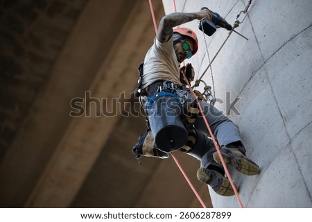 Rope access technician using a cordless drill while working on a concrete wall under a bridge
