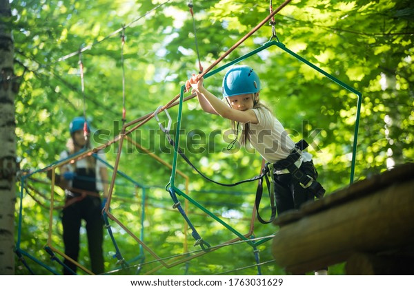 Rope adventure - a little girl walks on the rope\
bridge to the stand and holding by the ropes with her hands - her\
mother watching her