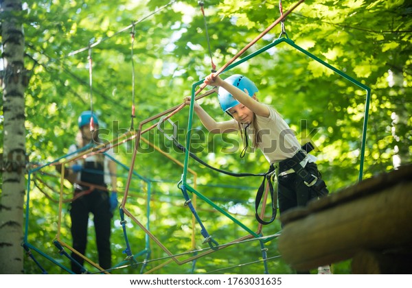 Rope\
adventure - a little girl walks on the rope bridge and holding by\
the ropes with her hands - her mother watching\
her